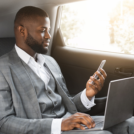 Man in a suite in the back of car looking at his phone while on his laptop
