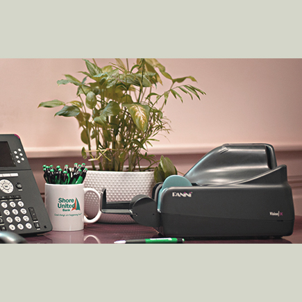 Desk with a phone, tape dispenser, plant, and Shore United Bank mug filled with pens